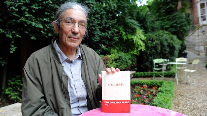 Assis dans un jardin, Boualem Sansal montre son livre « Rue Darwin ». 
