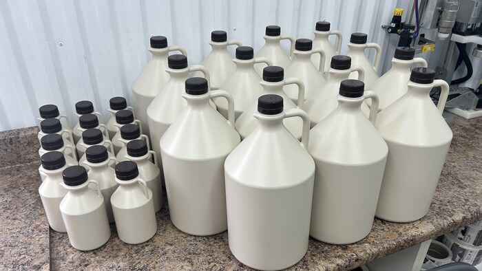 Des dizaines de bidons en plastique blanc cassé avec des bouchons noirs, de différentes grosseurs, sont posés sur un comptoir.