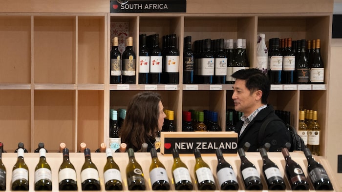 Un client et une employée, de profil, discutent dans un magasin d'alcool, entre des étalages de vins provenant d'Afrique du Sud et de Nouvelle-Zélande, tandis que les tablettes des vins des États-Unis ont été vidées de leur contenu.