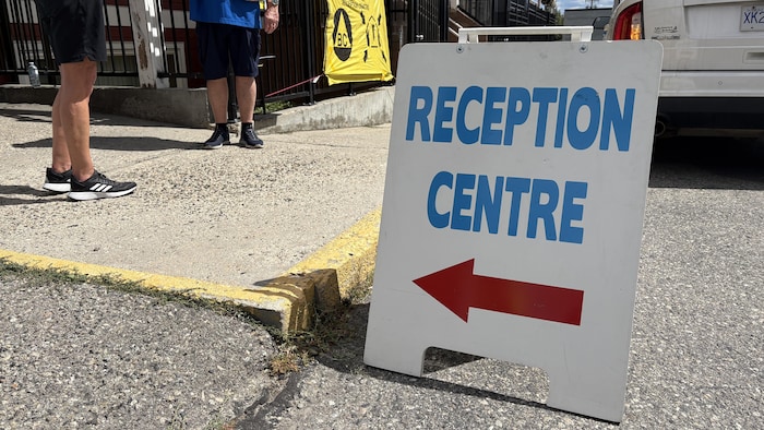 Un panneau blanc portant l’inscription « Reception Centre » en lettres bleues et une flèche rouge pointant vers la gauche est installé devant le centre d’accueil. Deux personnes et un véhicule blanc sont visibles à l’arrière-plan.