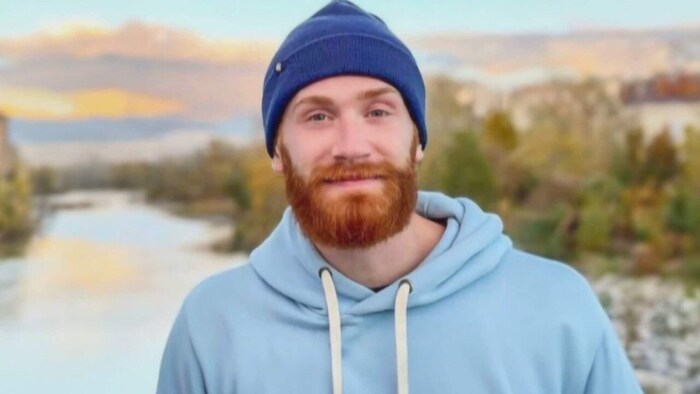 Un homme souriant, portant une barbe rousse et une tuque bleue, en automne devant un lac.