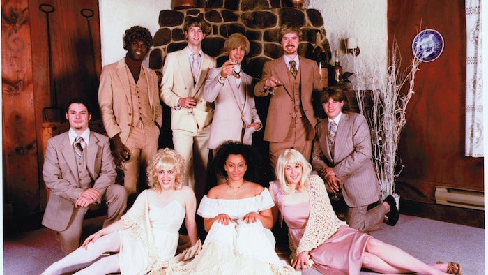 Neuf musiciens et musiciennes du groupe Bran Van 3000 prennent la pose, habillés à la mode des années 1970. 