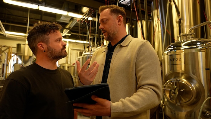 Deux personnes discutent des informations qu'elles lisent sur une tablette numérique, entourées par des cuves de brassage de bière.
