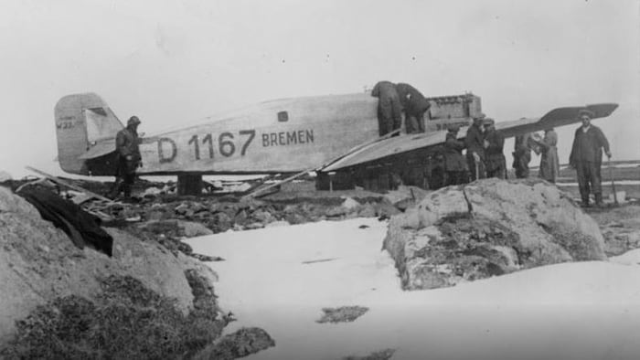 Le Bremen en difficulté sur l'île Greenly. Des hommes sont penchés au-dessus du cockpit et l'hélice a disparu. Quelques personnes s'affairent aussi autour de l'avion.