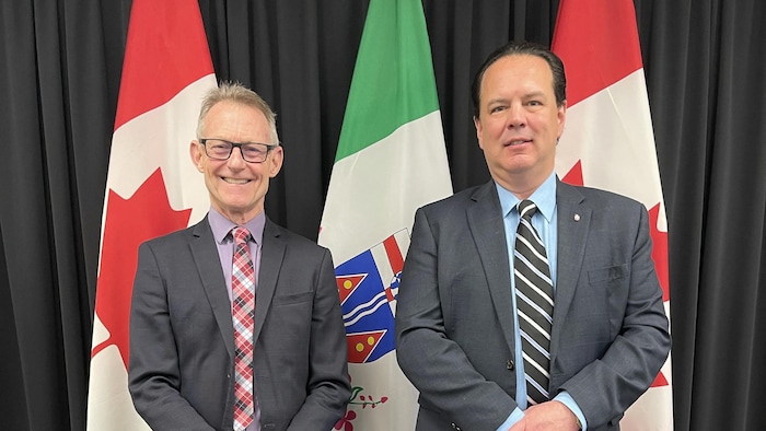 Brendan Hanley et Brad Cathers sont l'un à côté de l'autre devant trois drapeaux.