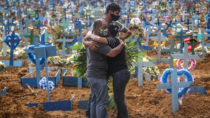 Deux personnes se serrent dans leurs bras, pendant qu'un autre regarde le nombre impressionnant de croix plantées dans le sol qui l'entourent.