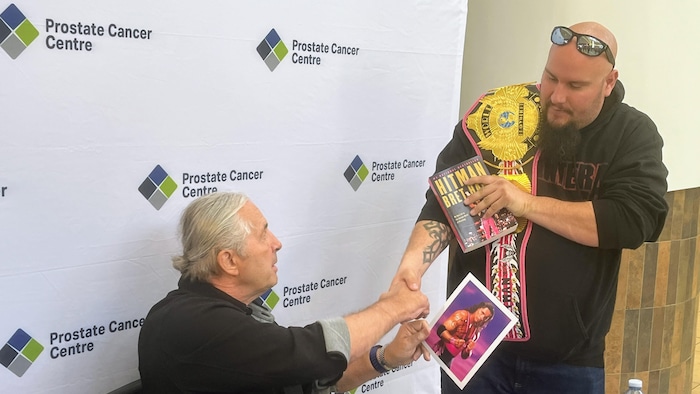L'ancien catcheur Bret « The Hitman » Hart, est assis à une table devant une murale du Prostate Cancer Centre et serre la main d'un partisan.