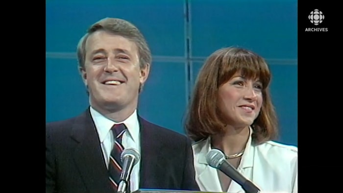 Brian et Mila Mulroney souriants, derrière des micros. 