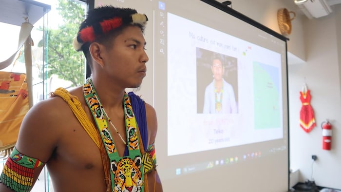 L'homme vêtu d'habits traditionnels autochtones se tient debout devant un écran où est projetée une présentation.