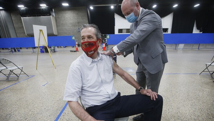 Le premier ministre, assis et masqué, reçoit le vaccin que lui administre Brent Roussin. 