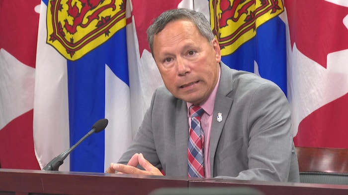 Assis en conférence de presse, le ministre joint les doigts de ses deux mains et grimace en parlant.