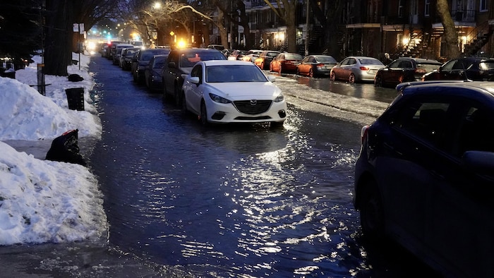 Une rue avec des maisons en rangée et beaucoup de voitures garées est inondée d'eau, dans la nuit.