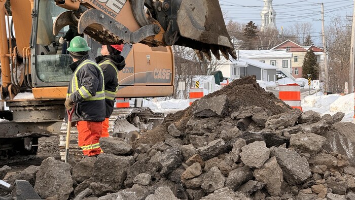Une pelleteuse, un tas de morceaux d'asphalte ainsi que des ouvriers avec des pelles dans une rue résidentielle.