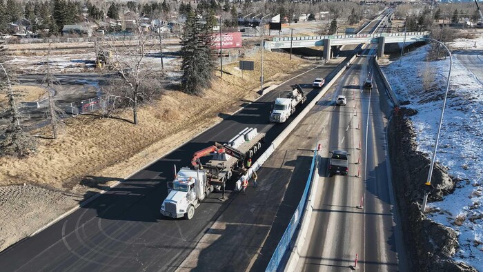 Vue aérienne d'un chantier routier où un camion à plateforme, muni d'un bras articulé, décharge des barrières de béton jersey le long d'une nouvelle voie asphaltée.
