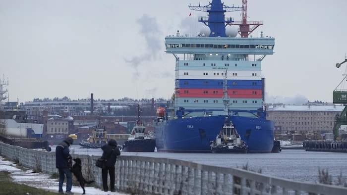 Le brise-glace Ural navigue dans un canal de Saint-Pétersbourg. Au loin, les édifices de la ville. Au premier plan, des piétons et un chien qui déambulent le long du canal.