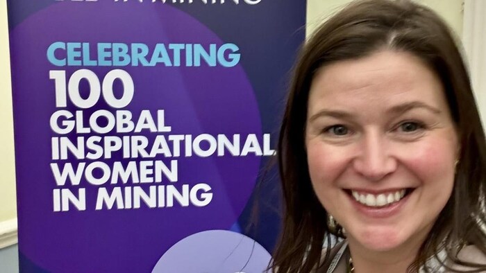 Une femme sourit devant une affiche qui célèbre 100 femmes inspirantes dans l'industrie minière.
