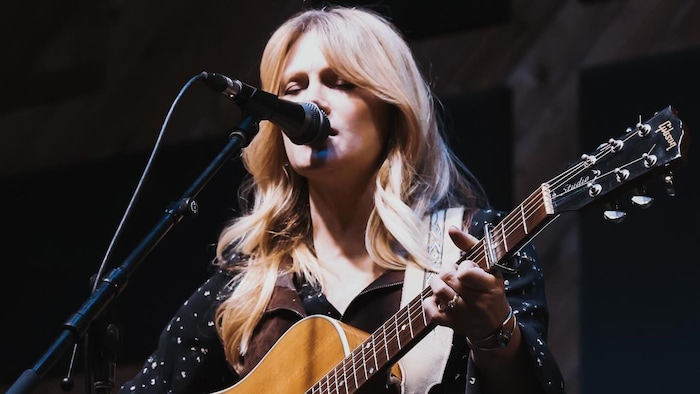 Une femme blonde chante dans un micro et tient une guitare. 