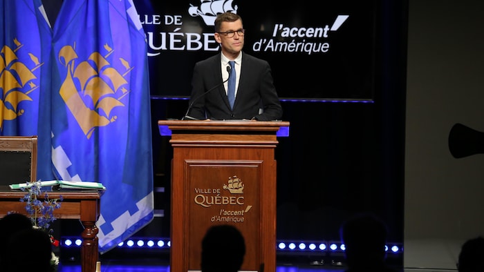 Bruno Marchand parle à un lutrin sur une scène avec la drapeau de la ville de Québec derrière lui.