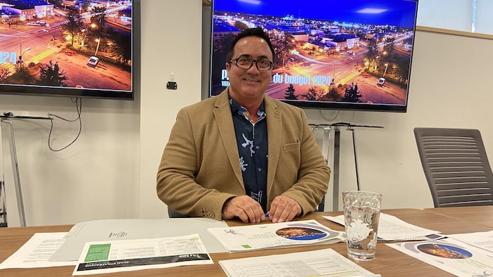 Yves Dubé, assis derrière une table couverte de documents, sourit à la caméra.