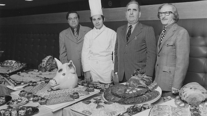 Quatre hommes se font prendre en photo devant une table garnie de nourriture (charcuteries, rôtis, aspics, etc.).