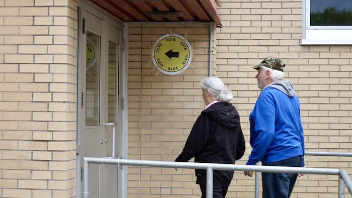 Deux personnes se dirigent vers l'entrée d'un bâtiment. Une affiche indique "vote" avec une flèche.