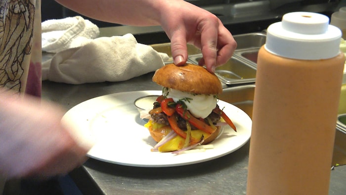 Une main potelée appuie sur la moitié supérieure d'un pain à hamburger, posée au-dessus d'une empilade de viande, poivrons et fromage fouetté.
