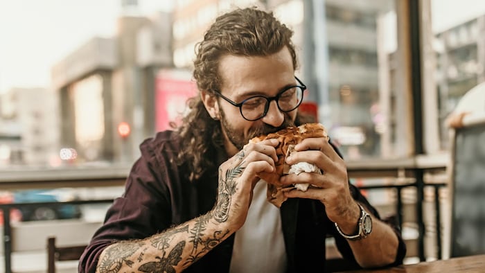 Un individu aux longs cheveux frisés, portant barbe et lunettes, est assis sur une terrasse et, les manches relevées, il met un burger dans sa bouche.