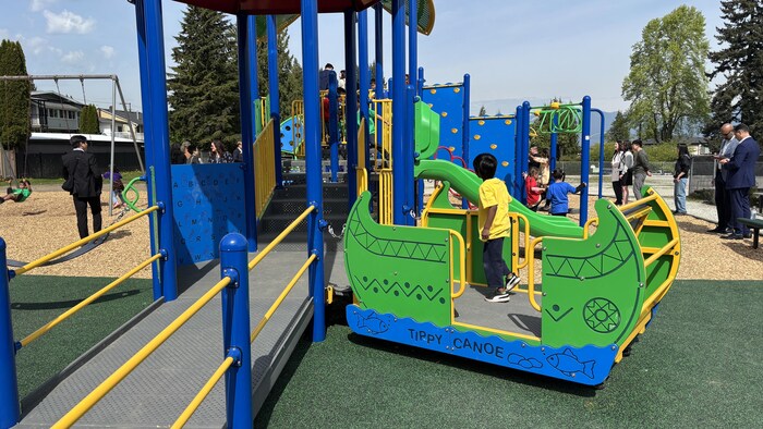 Un terrain de jeu pour enfant. Au milieu, un enfant dans une des parties du terrain de jeu. Au loin, d'autres enfants et des adultes autour. 
