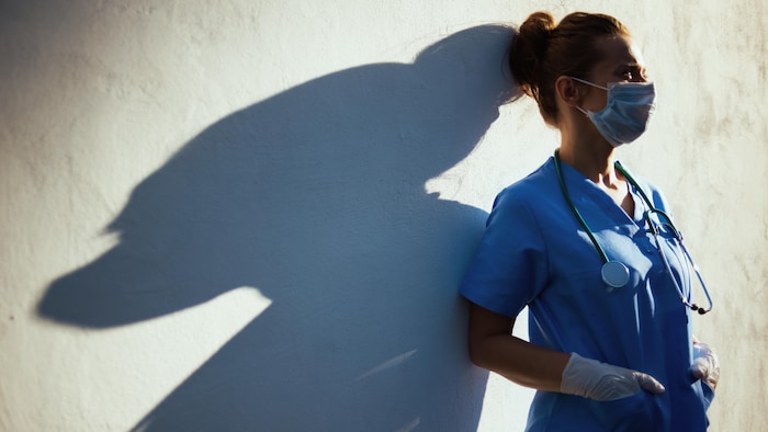 Une travailleuse de la santé accotée à un mur regarde au loin.