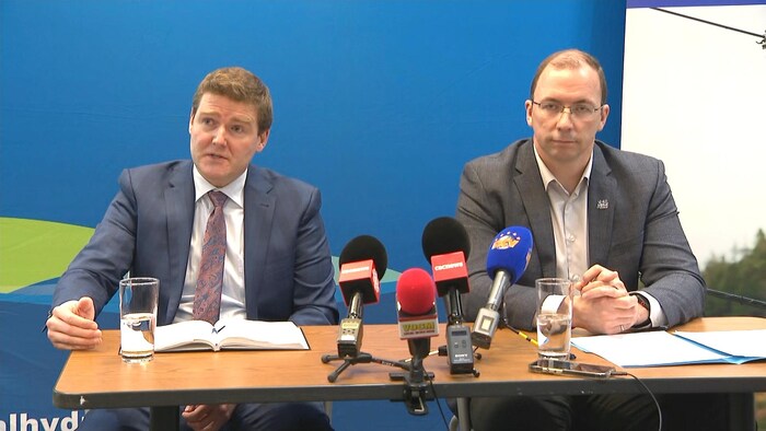 Deux hommes sont assis devant un mur bleu, à une table où sont posés des micros. L'homme de gauche parle.