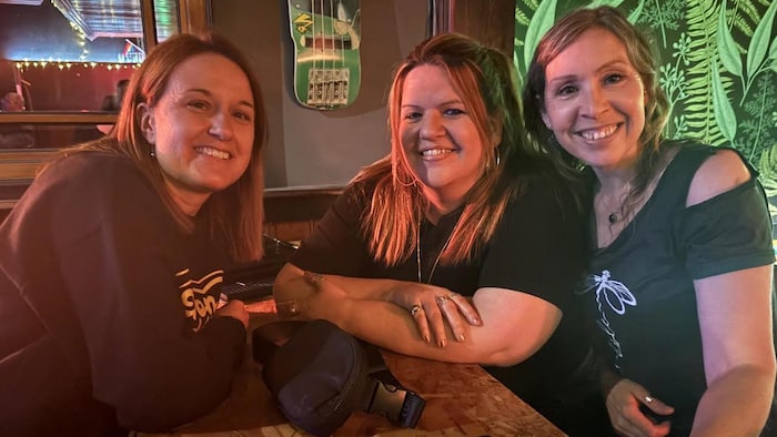 Trois femmes souriantes serrées les unes contre les autres, près d'un bar.