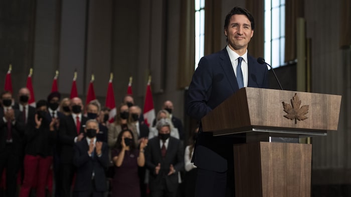 Justin Trudeau sourit devant un pupitre avec son nouveau gouvernement en arrière-plan.