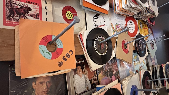 Des disques vinyle 45 tours sous pochette sont accrochés par leur centre à de longs clous sur un panneau en bois.