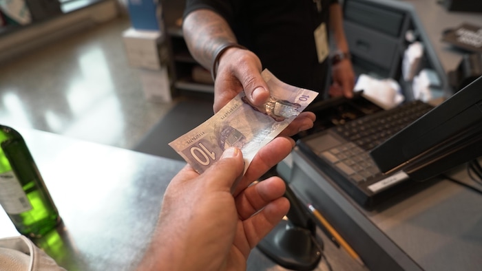 Un billet mauve sur lequel on peut lire le chiffre dix est tendu vers la caméra. Une main semble tendre le billet et une autre appartenant à une autre personne semble le prendre.