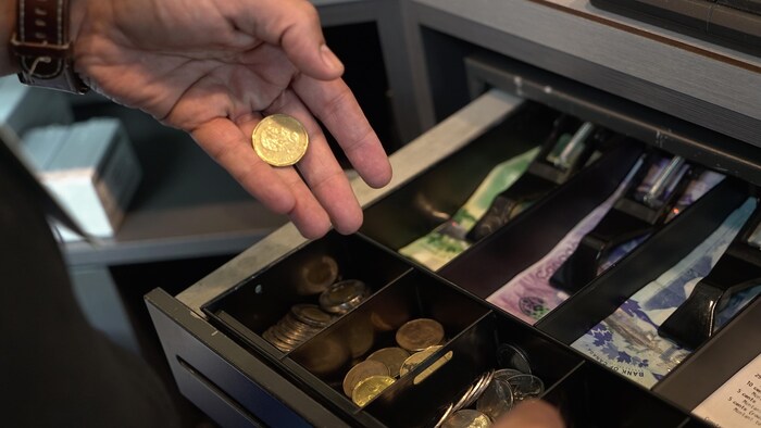 Une main, se trouvant au-dessus d'une caisse enregistreuse contenant des billets d'argent canadien et de la monnaie canadienne, tient une pièce de monnaie dorée.