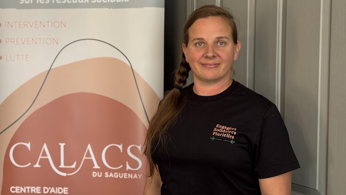 Une femme aux cheveux bruns tressés sur le côté et portant un t-shirt noir pose devant une bannière « CALACS DU SAGUENAY ».