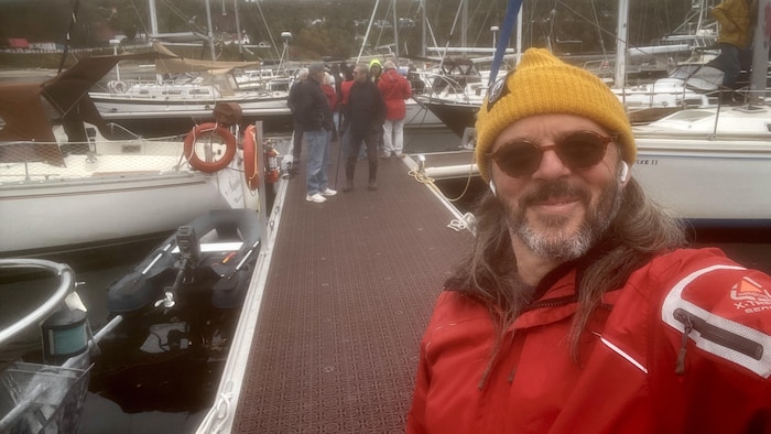 Un égoportrait pris par Éric Cyr. Il se tient sur le ponton flottant d'une marina, avec d'autres voiliers et des personnes à l'arrière-plan.