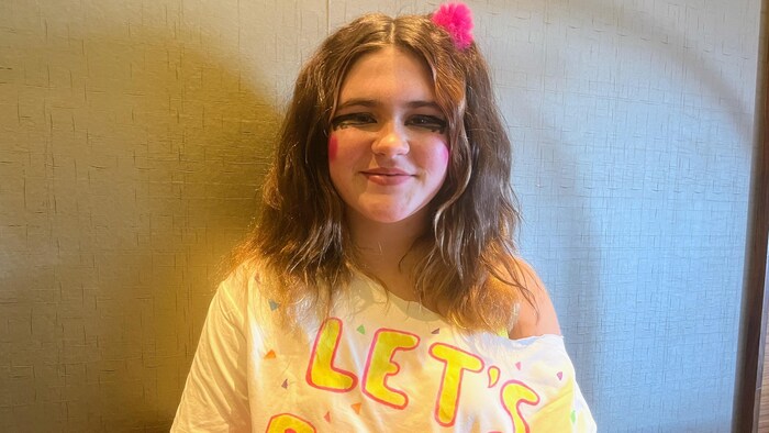 Une jeune fille vêtue d'un chandail blanc, d'accessoires rose fluo et de maquillage thématique tient une petite peluche brune dans ses bras.