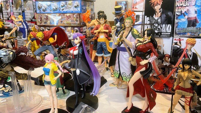 Une rangée de figurines de personnages de séries d'animation japonaises, dont plusieurs de la franchise One Piece, est exposée sur une table devant leurs boîtes d'emballage d'origine.