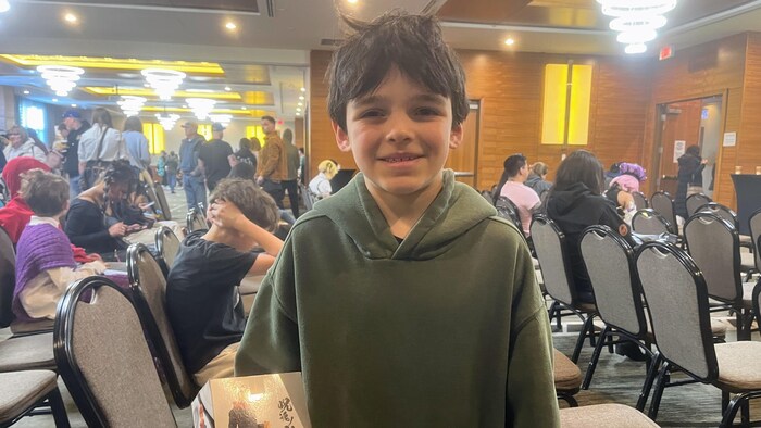 Un jeune garçon sourit à la caméra tout en tenant fièrement une boîte contenant une figurine de collection dans une salle de conférence remplie de chaises.