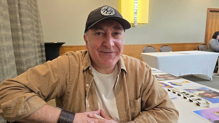 L'acteur de doublage Peter Kelamis sourit devant une table garnie de photos dédicacées et de produits dérivés lors d'un festival