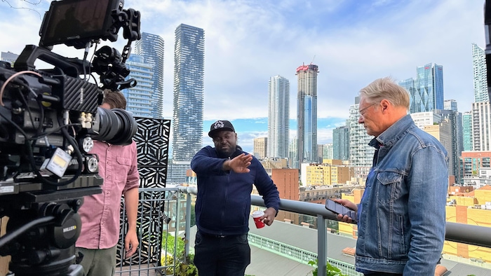 Un caméraman se tient derrière sa caméra, le réalisateur pointe du doigt une direction et un journaliste se tient en face de lui, un téléphone à la main. En arrière-plan, on voit la ville de Toronto avec ses gratte-ciel.