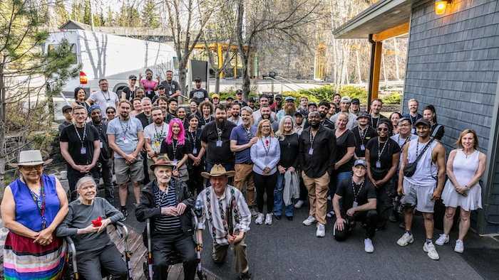 Une foule, pour la plupart composée de jeunes, pose pour une photo de groupe à l'extérieur d'un bâtiment en bois. 