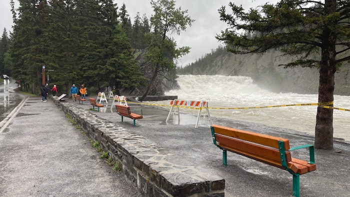 Un point de vue en bordure d'une rivière en crue avec des barrières de sécurité et du ruban jaune de signalisation bloquent l'accès à la berge en raison du fort courant de l'eau tumultueuse. 