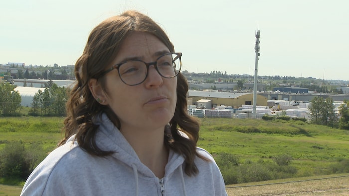 Portrait en extérieur d'une femme aux cheveux bruns mi-longs portant des lunettes et un chandail à capuchon gris, regardant hors champ lors d'une entrevue.