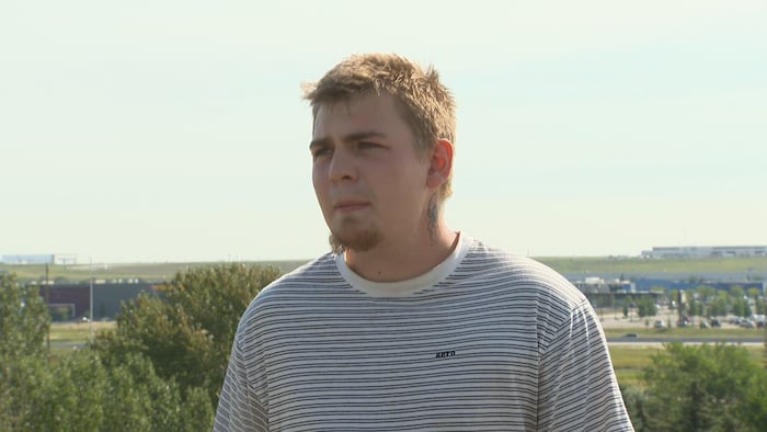 Portrait en extérieur d'un jeune homme aux cheveux courts avec une barbichette, un tatouage sur le côté gauche du cou et un t-shirt blanc à rayures foncées, regardant hors champ pendant une entrevue.