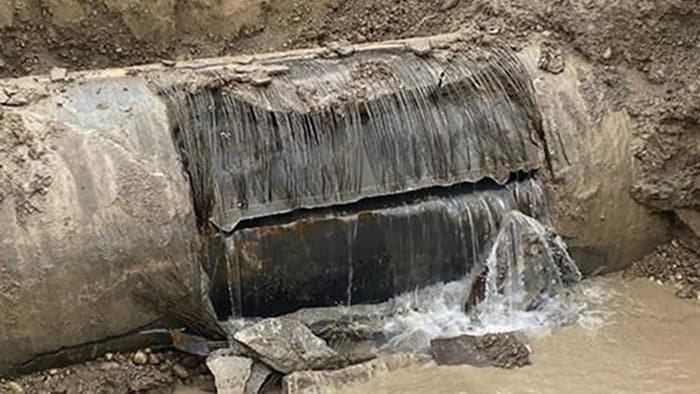 Une importante fuite d'eau jaillit d'une large fissure horizontale dans une conduite souterraine de grand diamètre mise au jour dans une tranchée d'excavation.