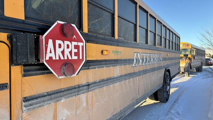 Gros plan sur le flanc gauche d'un autobus scolaire jaune marqué « INTERBUS ». Le panneau d'arrêt octogonal rouge avec l'inscription « ARRÊT » est visible au premier plan. Les véhicules circulent sur un chemin enneigé sous un ciel clair.