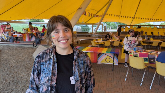 Portrait d'une personne avec un petit drapeau arc-en-ciel peint sur la joue, sous la tente jaune.