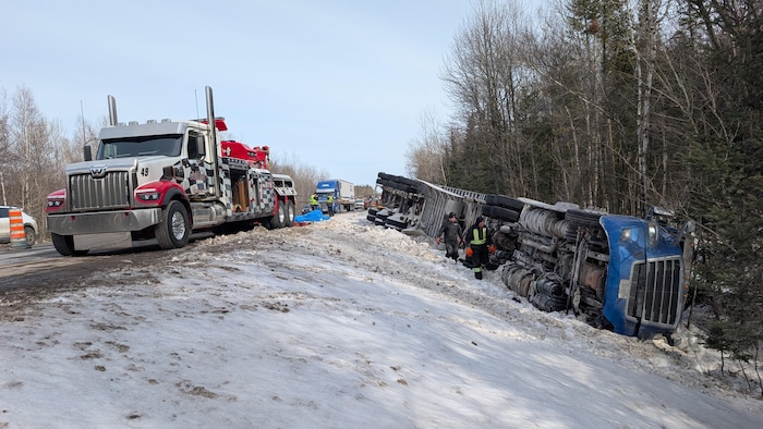 Un camion poids lourd est renversé aux abords d’une autoroute et d’un boisé. Deux autres camions se trouvent sur l’autoroute.
